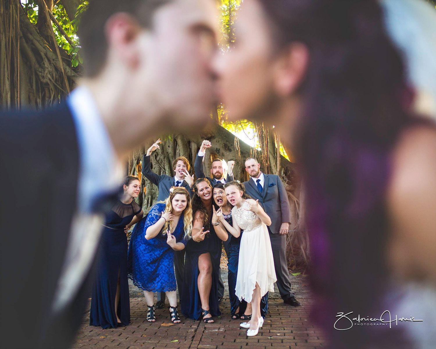 Wedding guests react with joy and laughter while gathered on a garden path framed by blurred foreground figures.