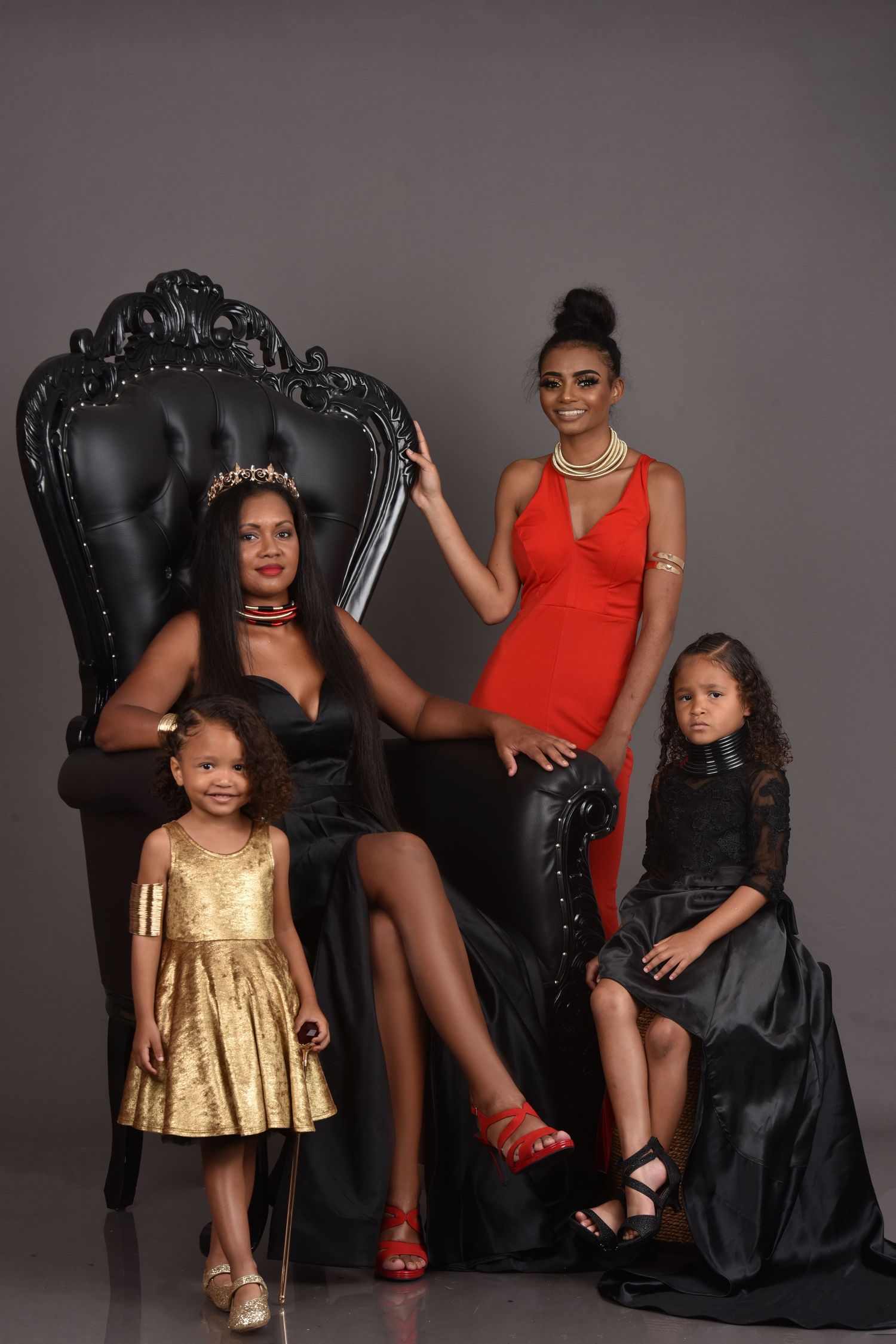 Four elegant women and girls in formal black, gold, and red attire pose with a ornate black throne chair in a studio.