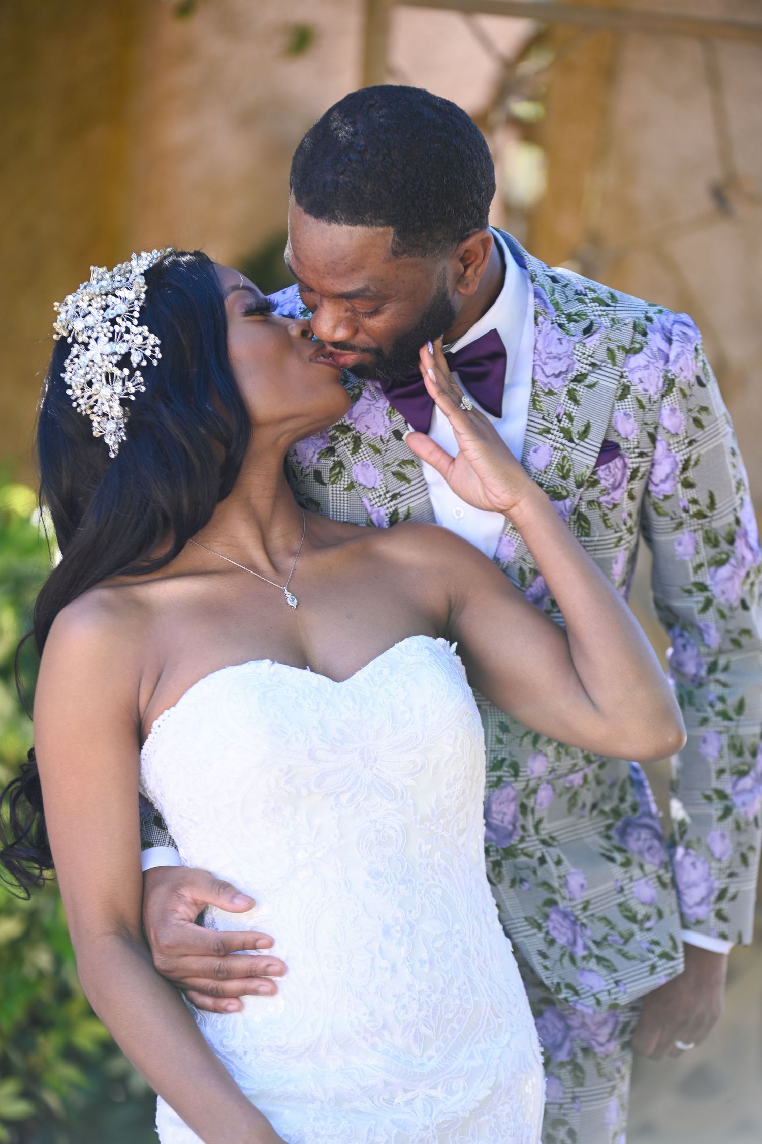 A bride in a white lace gown and groom in a floral suit share a romantic kiss at their wedding ceremony.