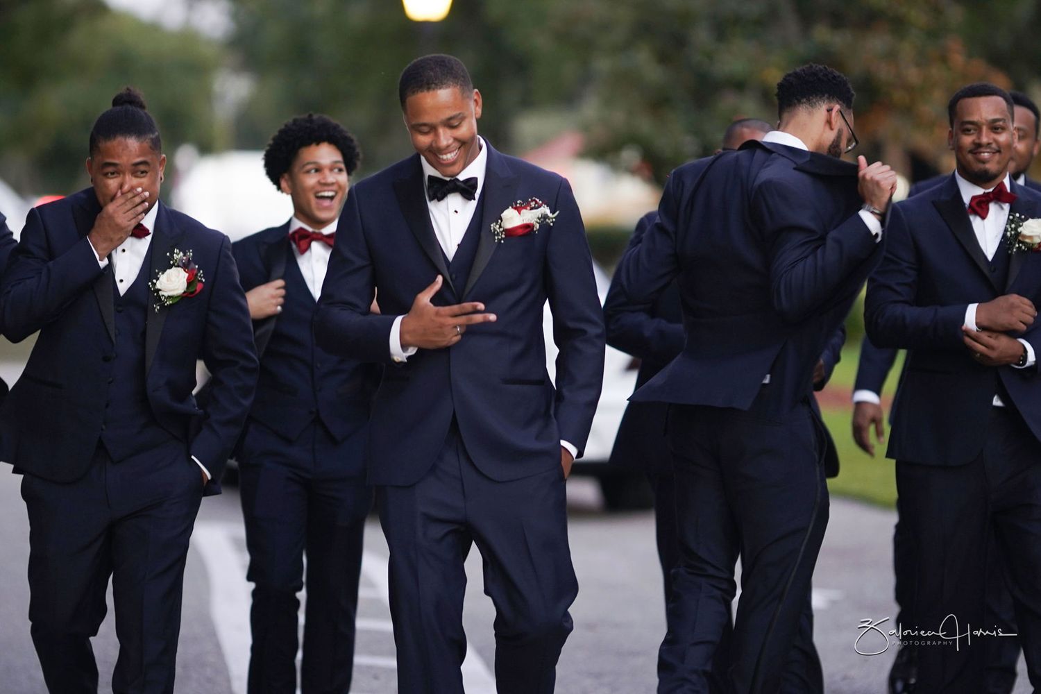 Groomsmen in black suits and bow ties laugh and smile together while looking at their phones during a wedding.