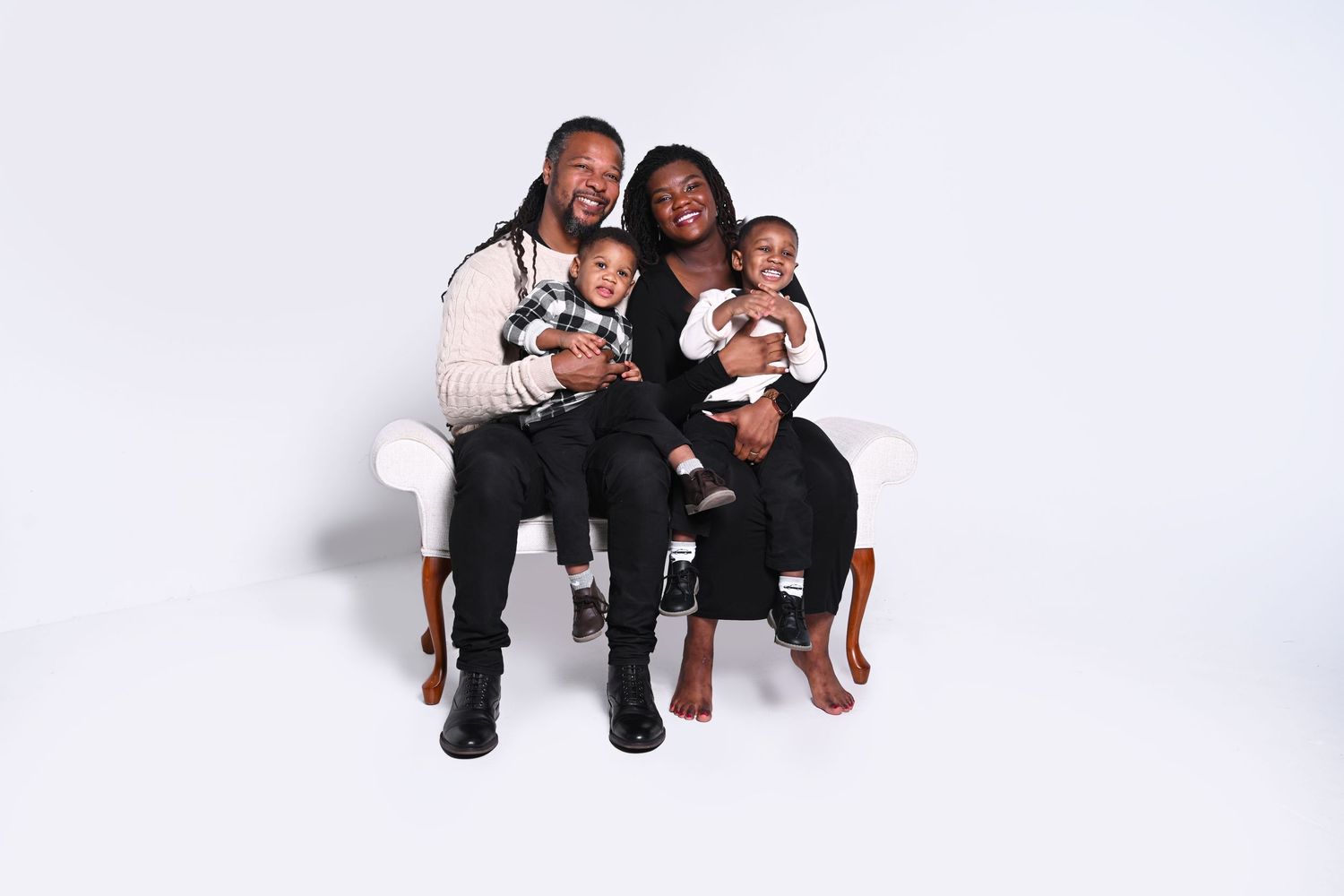 A joyful family of four poses on a white bench, with two parents holding their young children against a white background.
