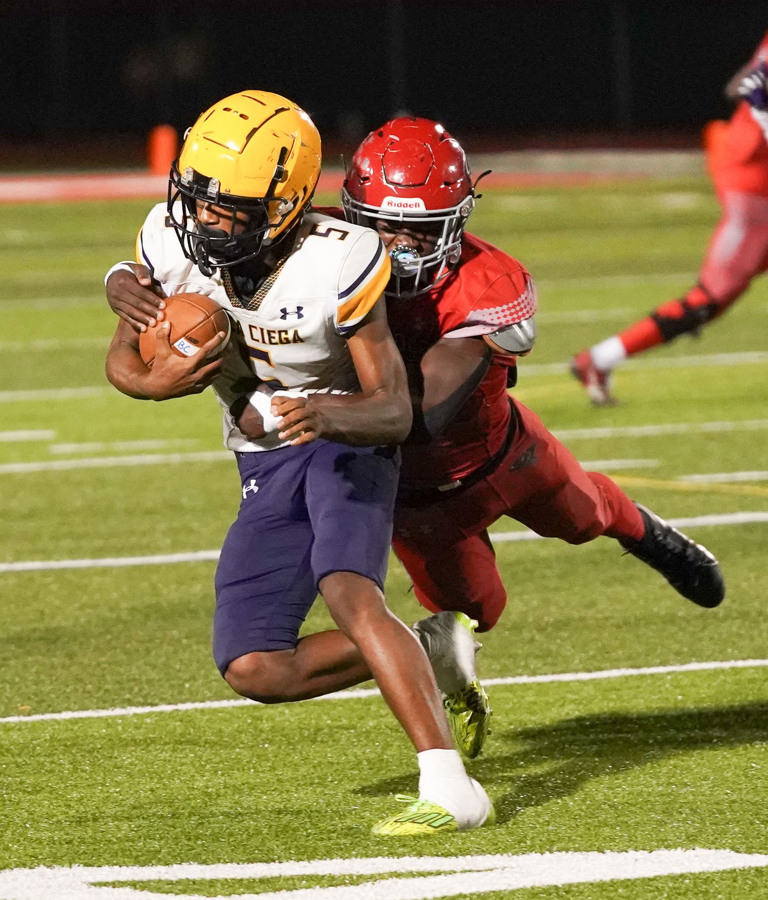 High school football player in gold uniform carries ball while red-jersyed defender attempts tackle during night game.