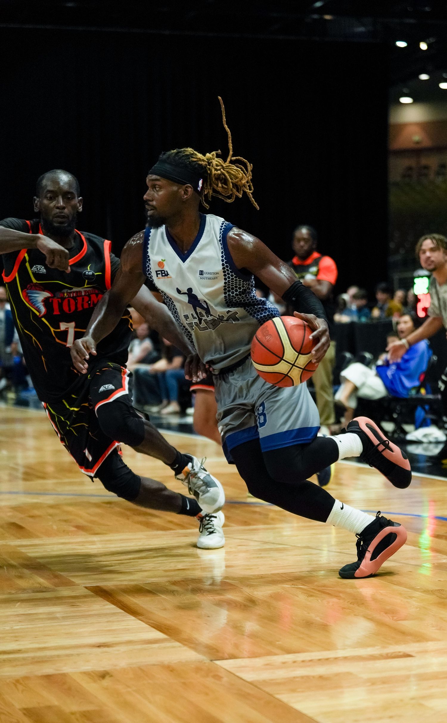 Basketball player with dreadlocks drives to the basket past a defender during an indoor game.