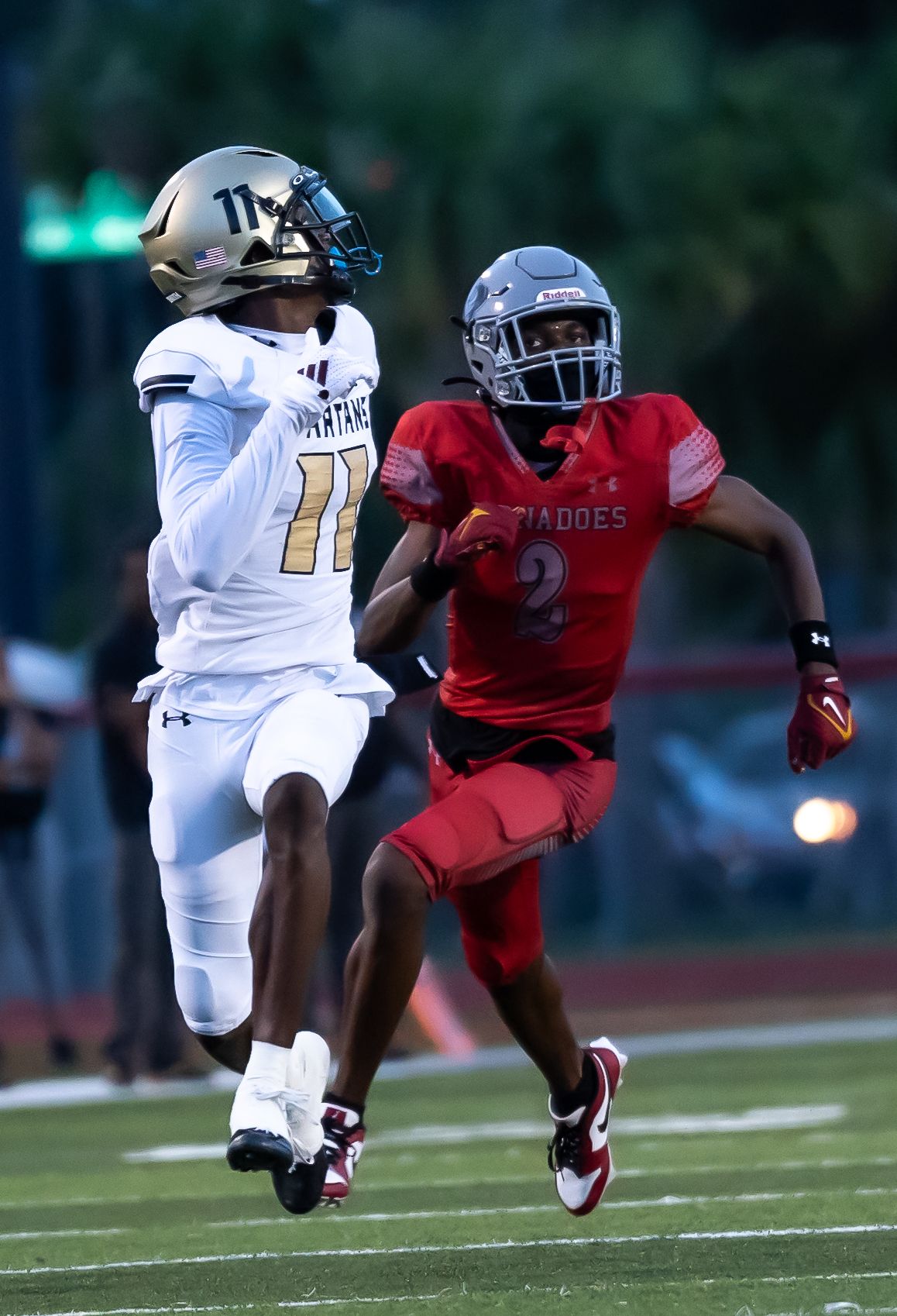 Two high school football players compete during a game, one in white uniform #11 and another in red #2.