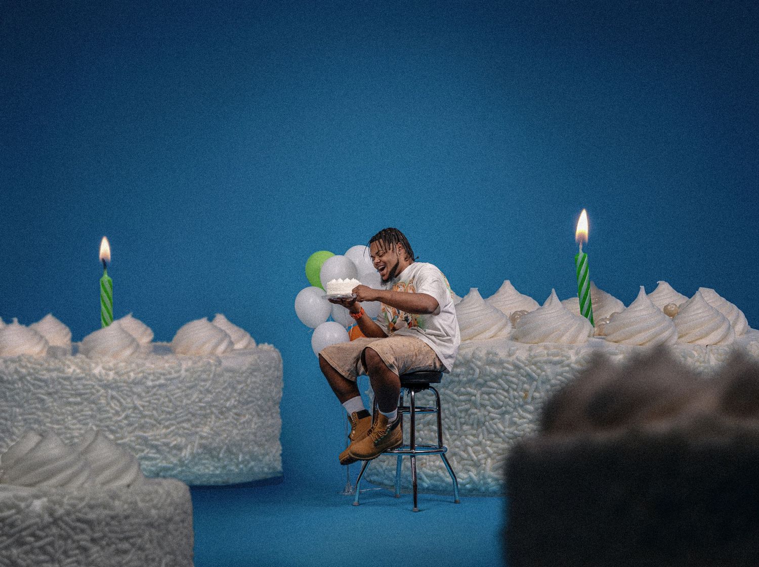 Man sitting alone between giant birthday cakes with lit candles and balloons against a blue background.