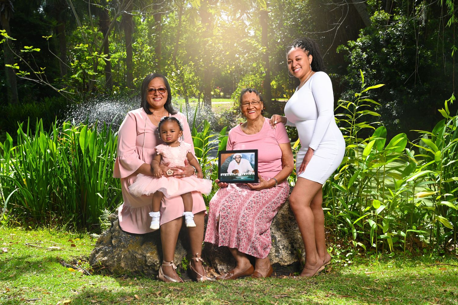 Four generations of women pose outdoors in a lush green garden, one holding a framed photo of a family member.
