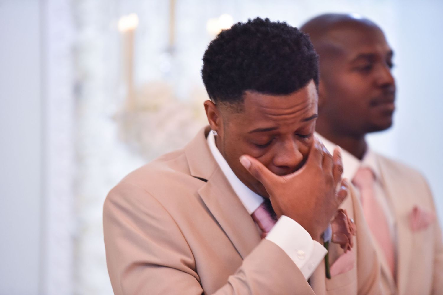Emotional groom in blush pink suit wiping tears at wedding ceremony with groomsman standing behind him.