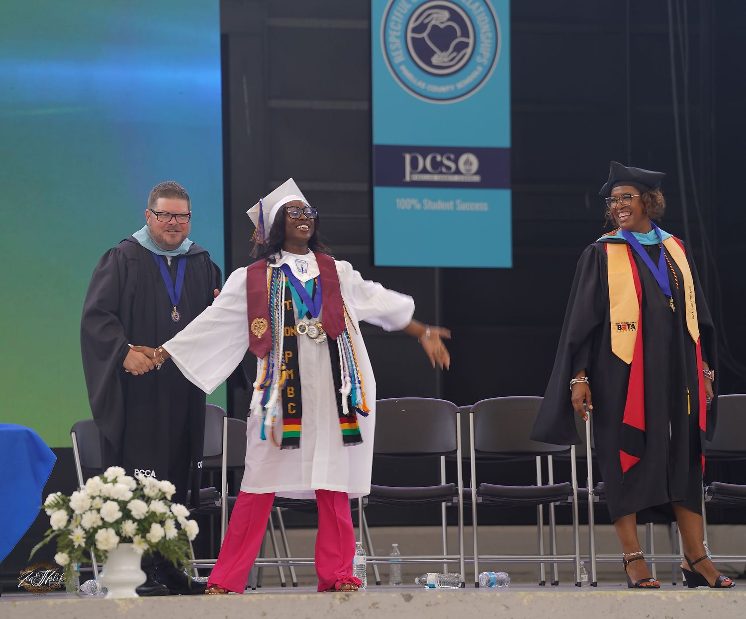 Graduates in caps and gowns walk across stage during commencement ceremony with white flowers on display in Tampa Florida.