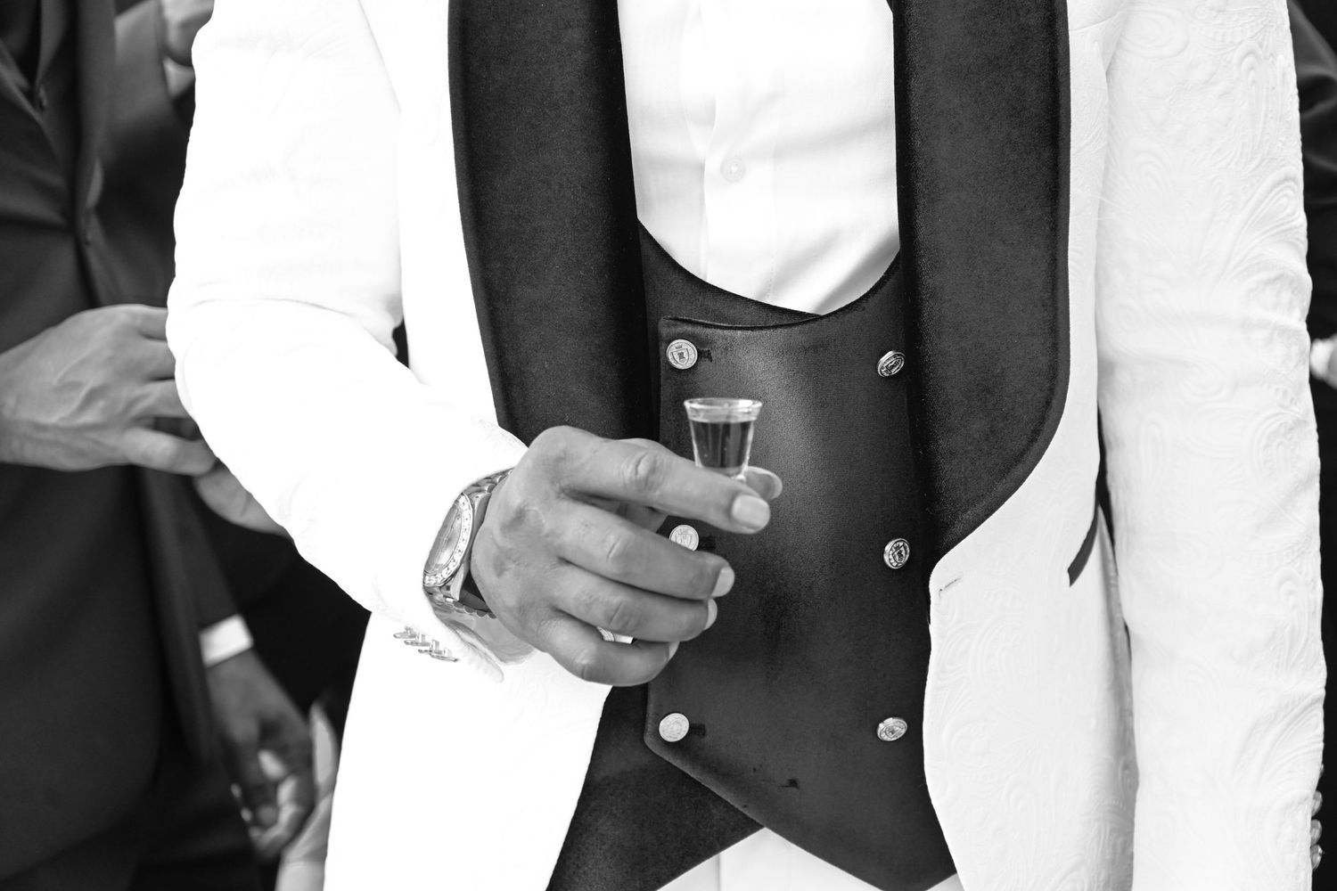 Man in white tuxedo jacket and dark vest holding a shot glass at a formal event, black and white photo.