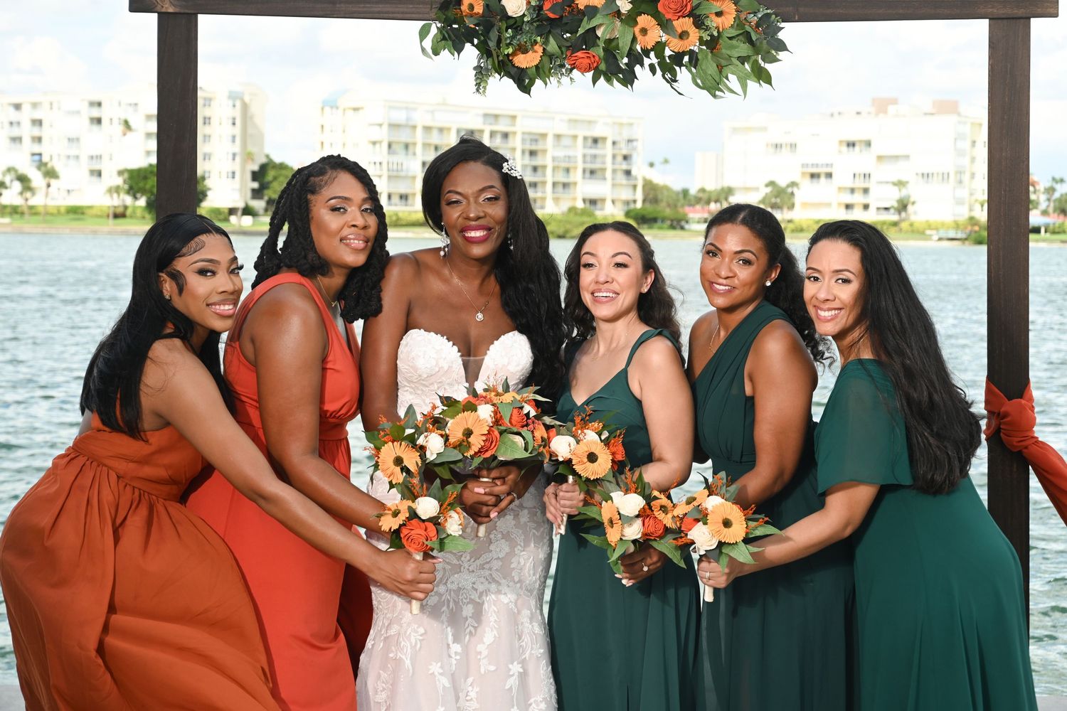 Wedding party in emerald green and rust orange dresses posing by waterfront.