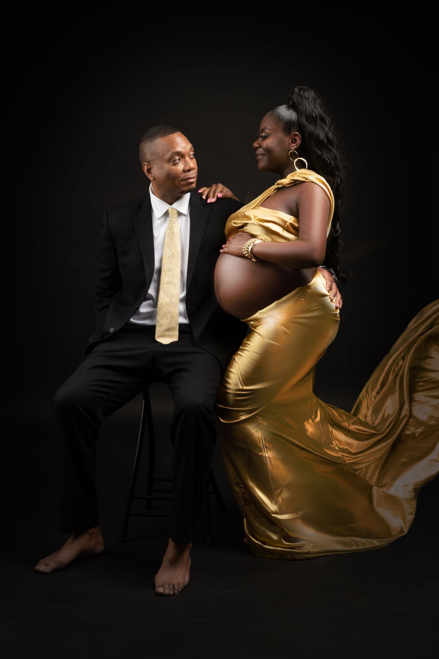 Elegant maternity session with flowing gold dress and formal attire against dark backdrop.