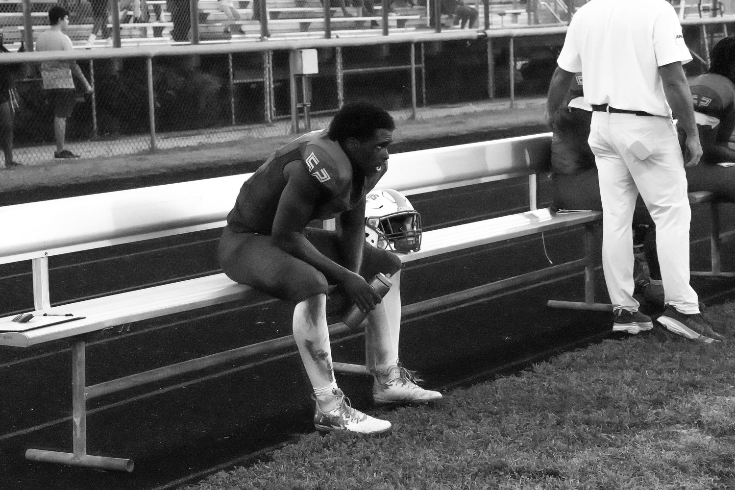 Black and white photo of a football player sitting alone on bleachers, holding a helmet on the sidelines.
