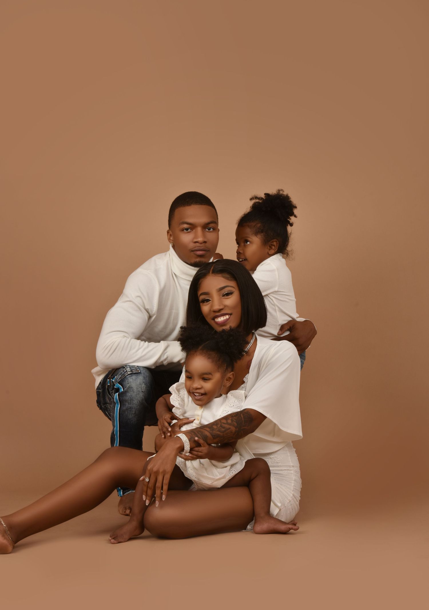 A family of four in white outfits poses together against a warm beige background in a professional studio portrait.