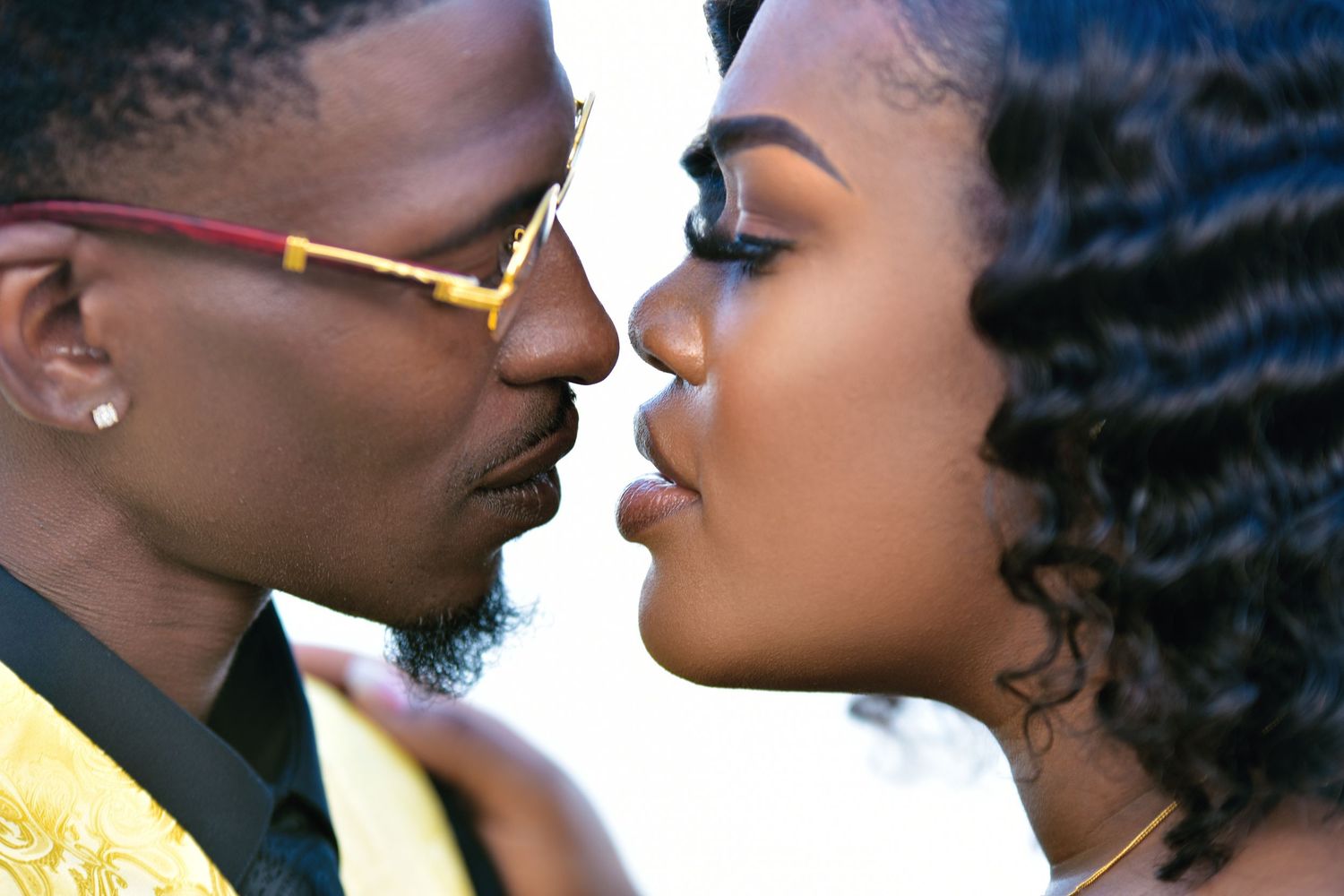 Close-up of couple nearly kissing, man wearing gold-framed glasses and gold patterned jacket.