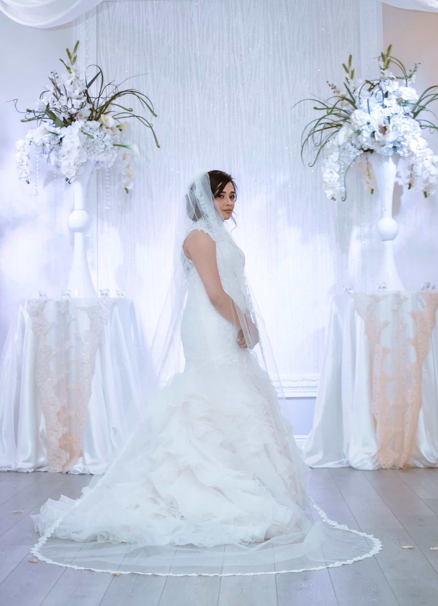 A bride in a flowing white wedding gown poses gracefully in a light-filled room with elegant floral arrangements.