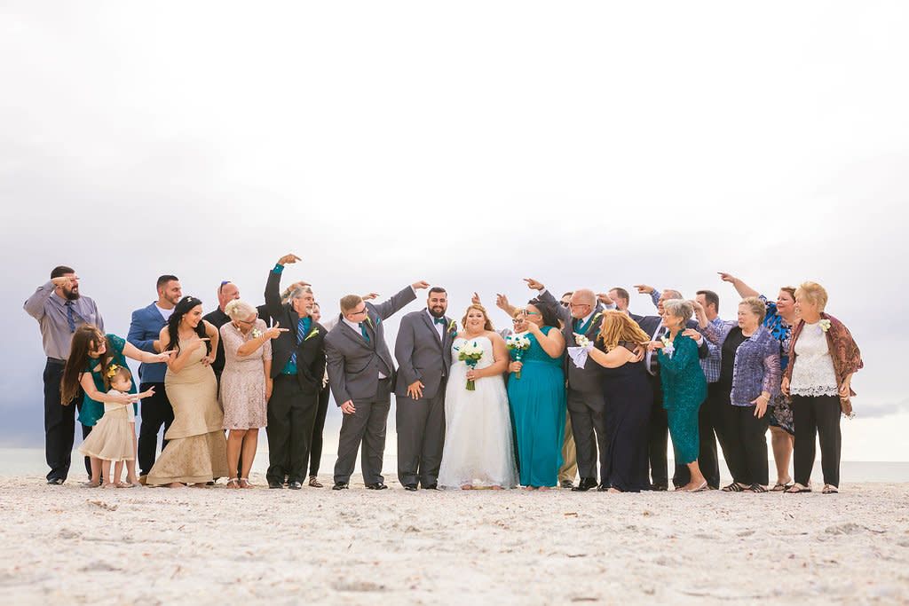 Large wedding party group poses together on white sandy beach during destination ceremony.