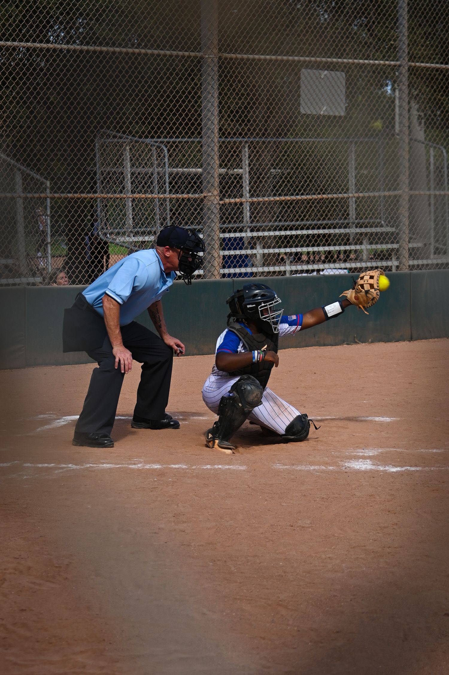 Softball catcher in full gear catches a pitch at home plate while the umpire watches during a youth softball game.