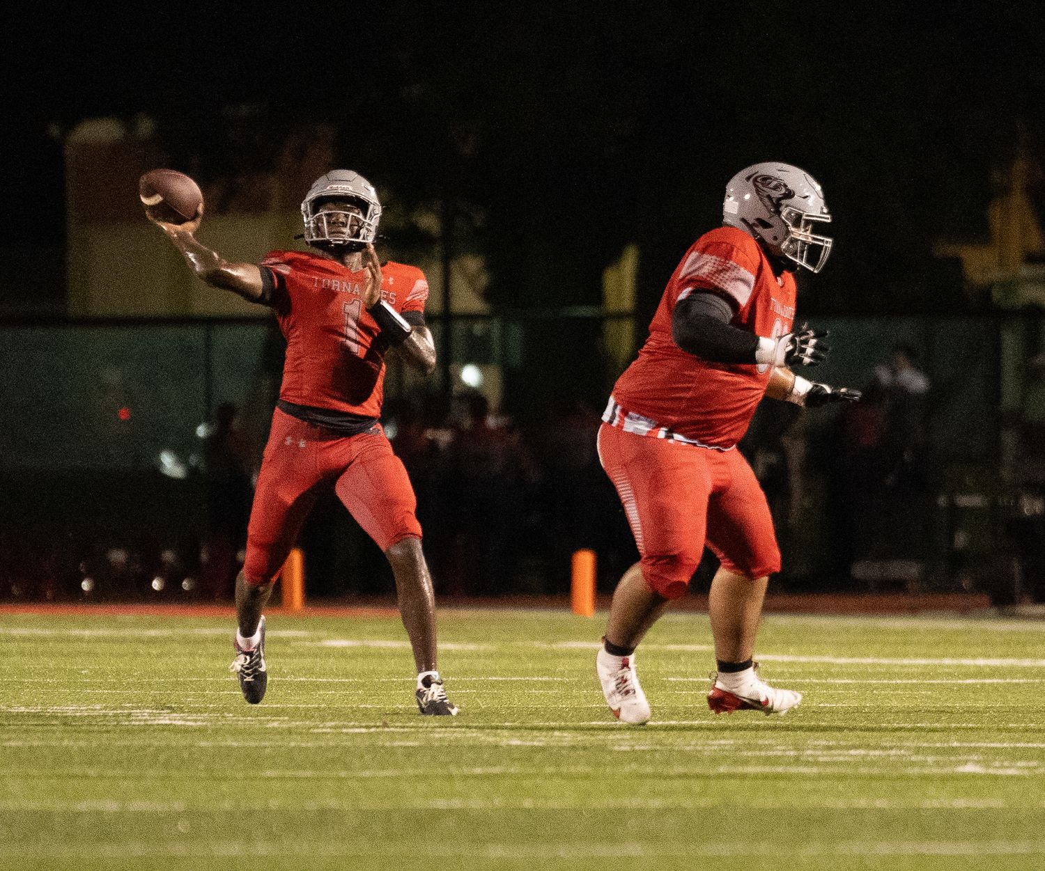 Two football players in red uniforms on a lit field, with the quarterback preparing to throw the ball.