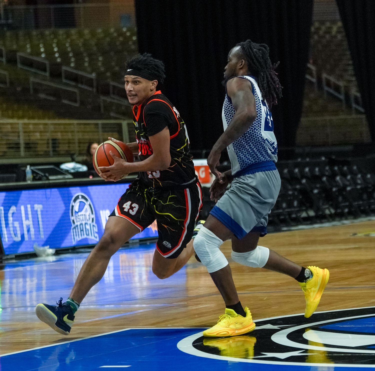 Basketball player #43 in black uniform drives to the basket past a defender in blue during an indoor game.