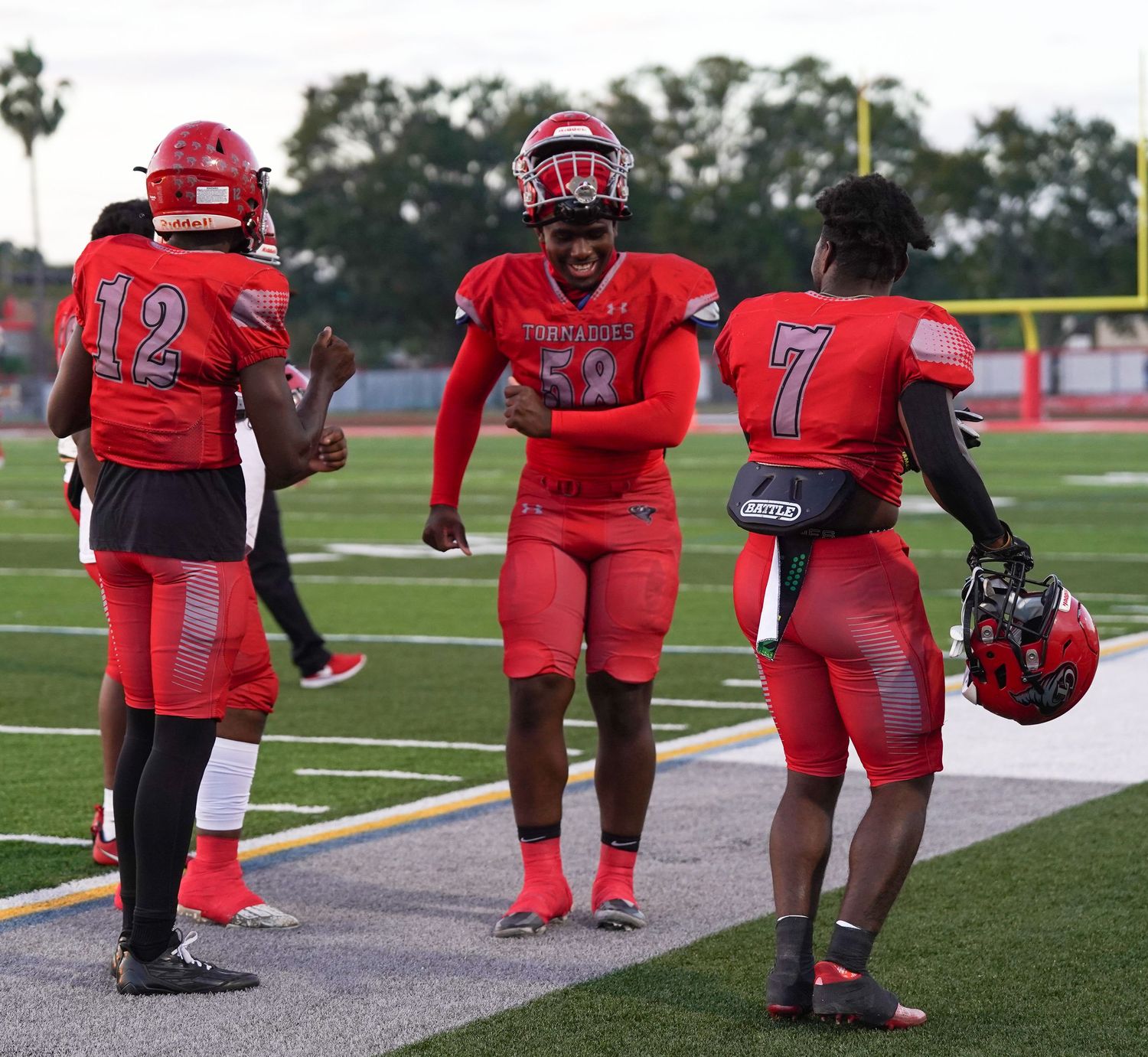 Three high school football players in red uniforms celebrate on the sideline during a game.