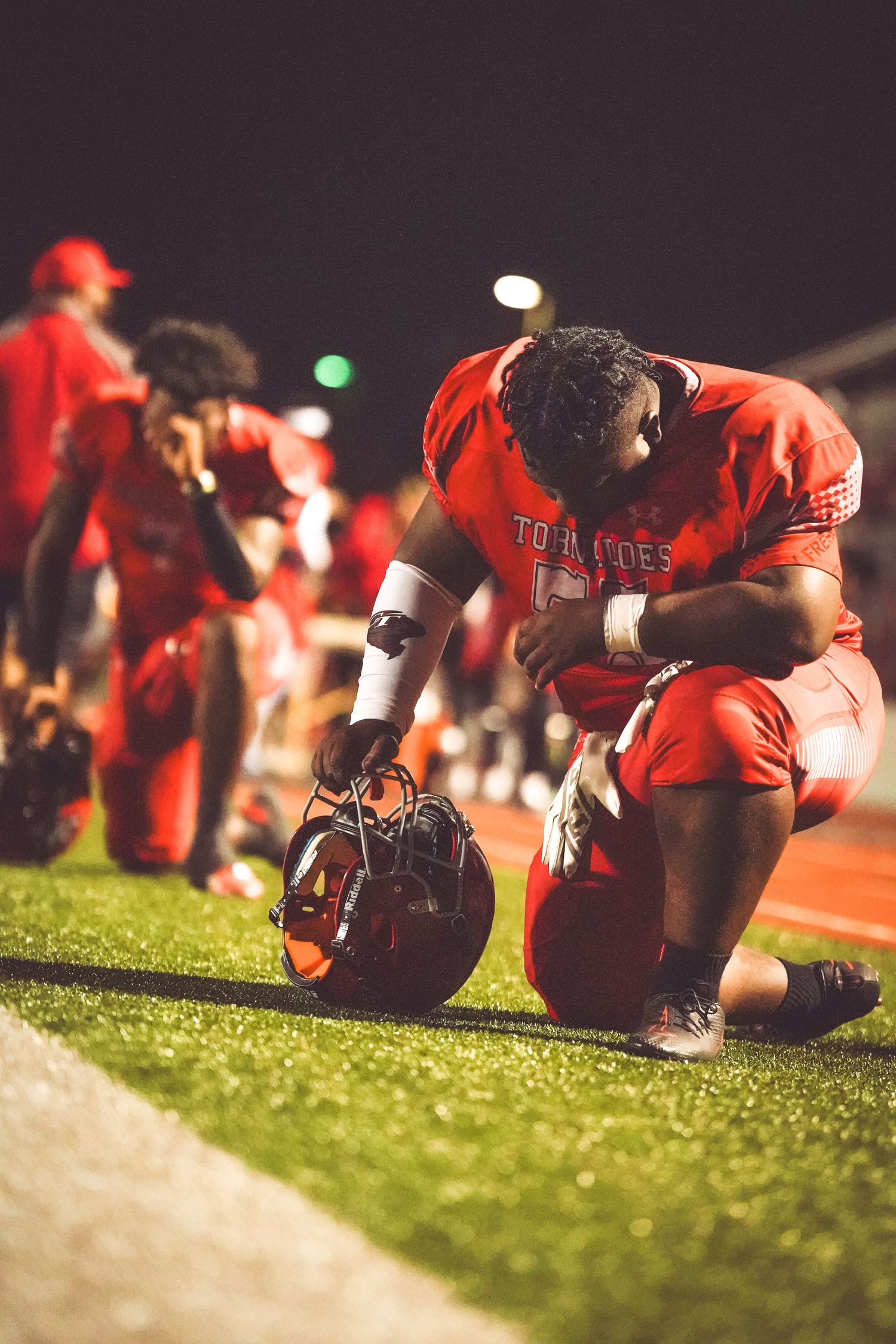 Football player in red Tornadoes uniform kneels on field holding helmet during nighttime game.
