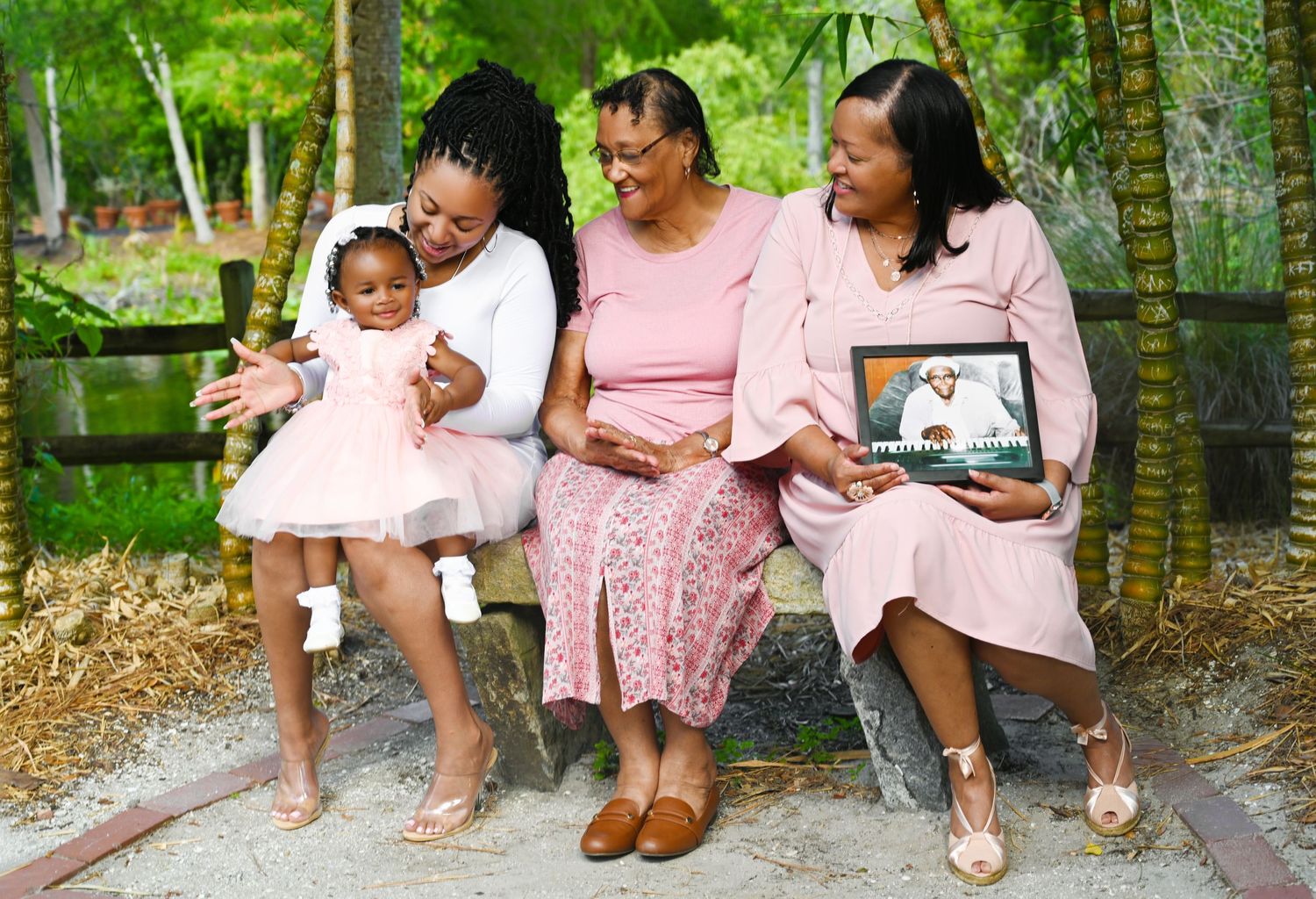 Four generations of women in pink outfits sit on a bench outdoors, sharing a joyful moment with a laptop photo.