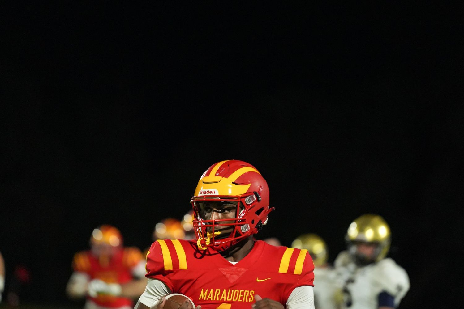 Marauders football player in red and yellow uniform holds ball under Friday night lights during a competitive game.