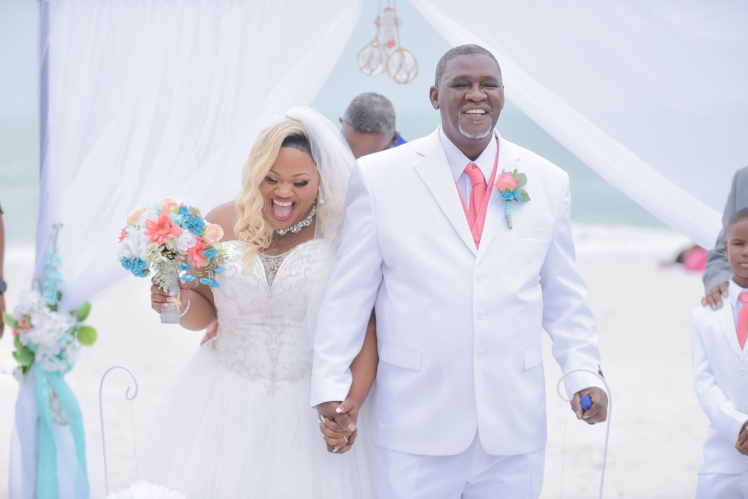 Happy newlyweds walk hand-in-hand on a beach wedding ceremony, bride in white gown holding coral bouquet.