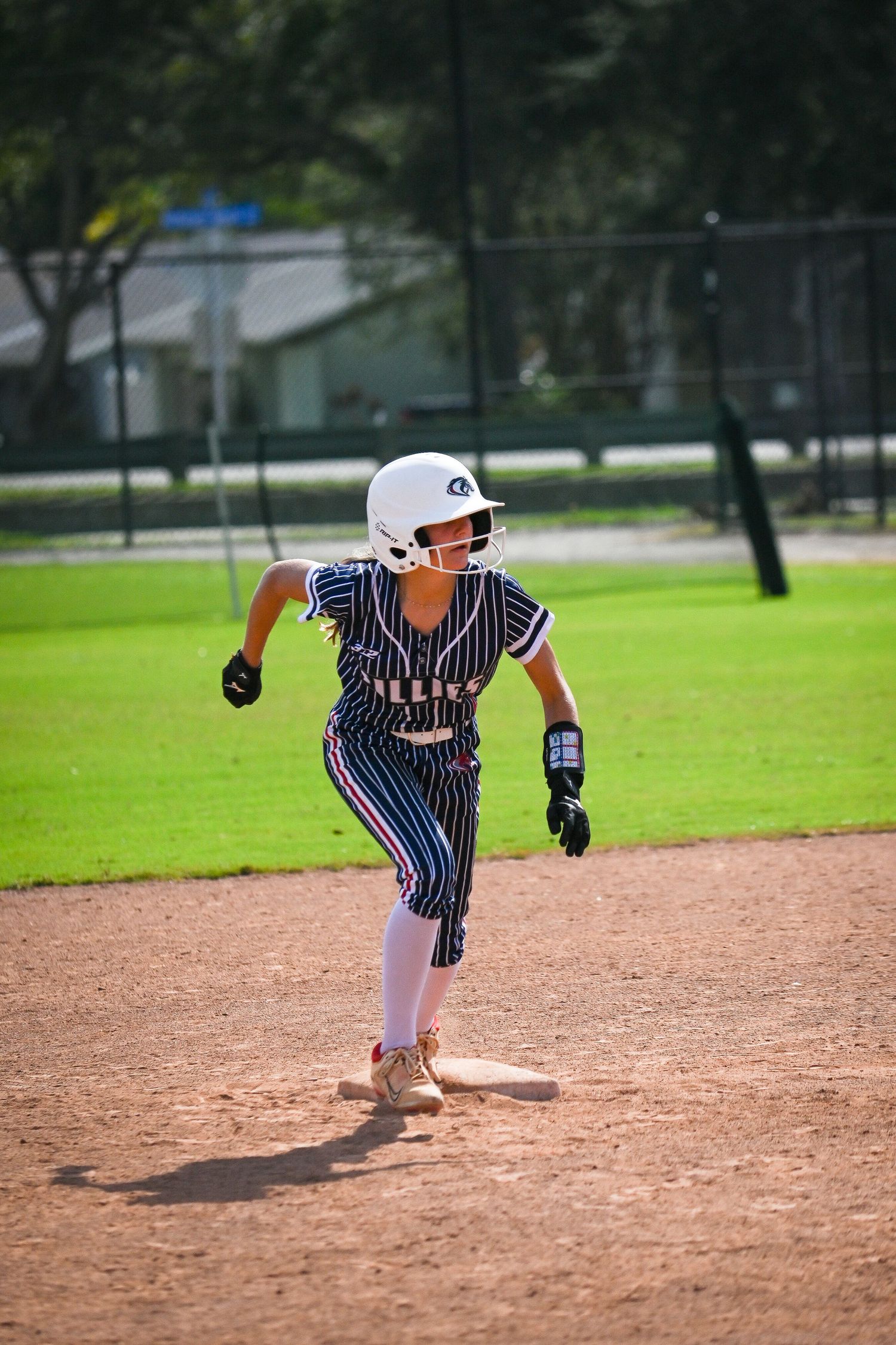 Young softball player in pinstripe uniform running bases during a sunny daytime game on a dirt infield.