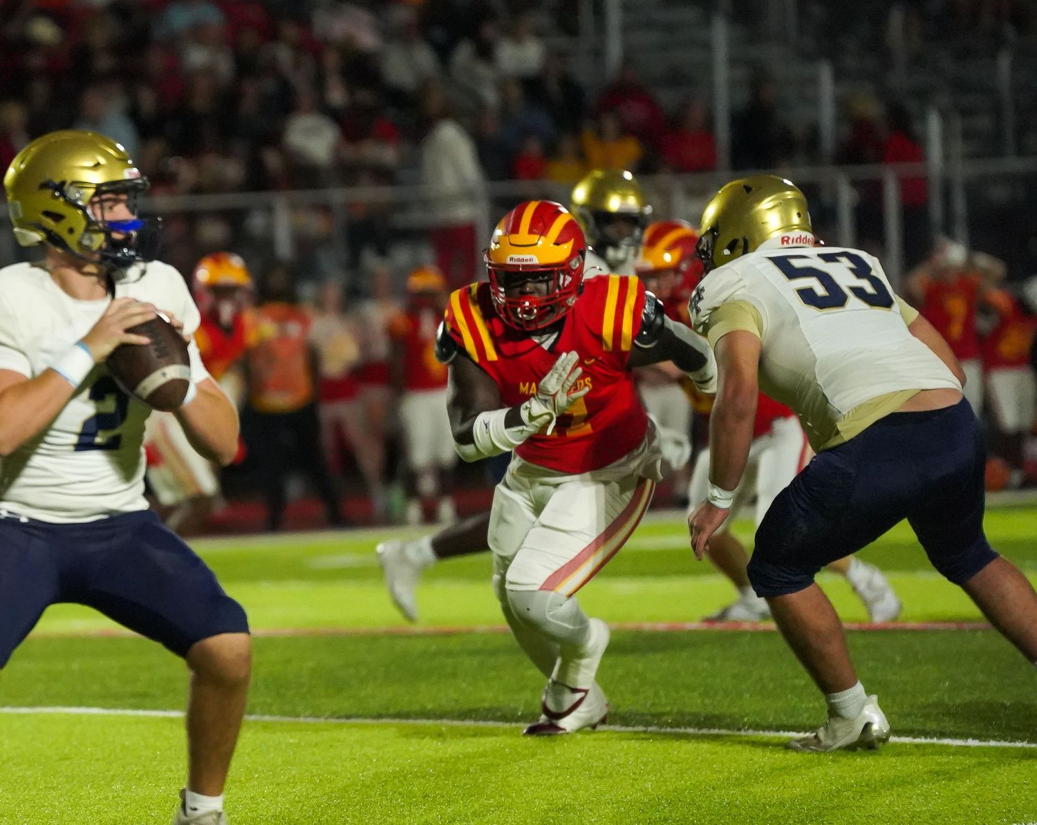 High school football player in red uniform breaks through defenders in navy jerseys during a night game.