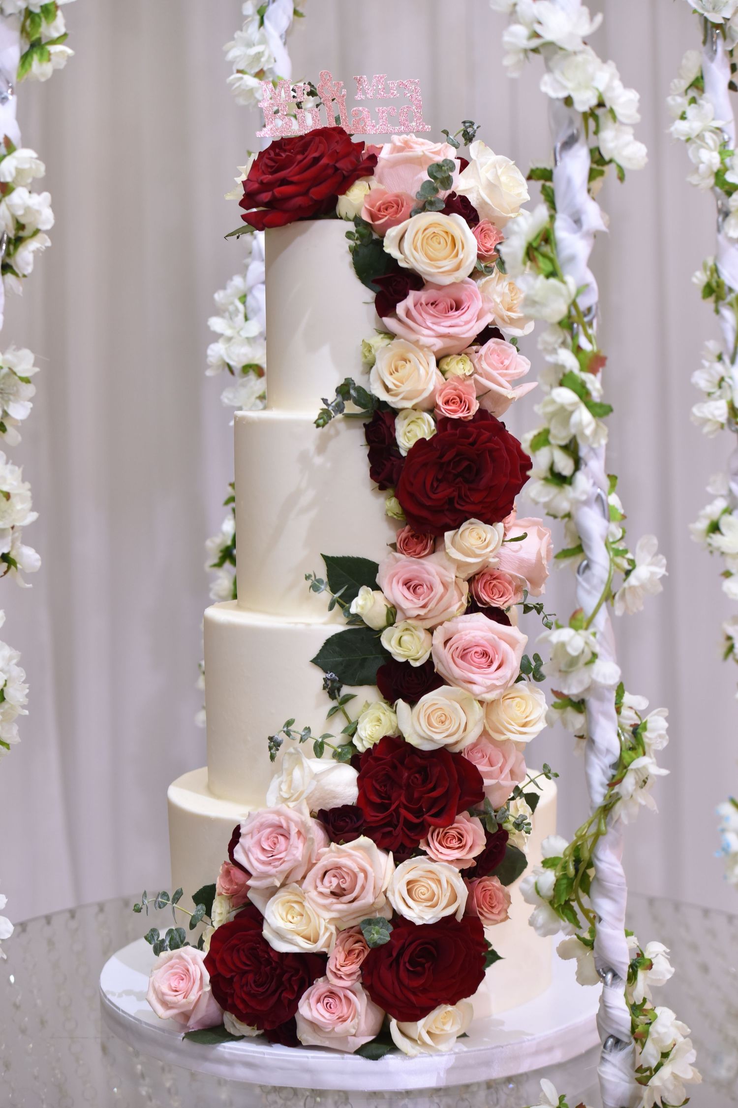 Elegant five-tier white wedding cake adorned with cascading red and pink roses and greenery.