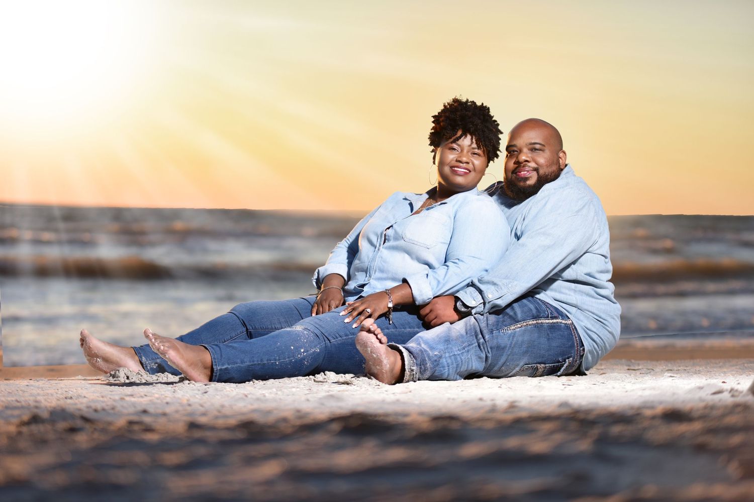 A couple sits together on a sandy beach at sunset, smiling warmly in matching denim outfits.