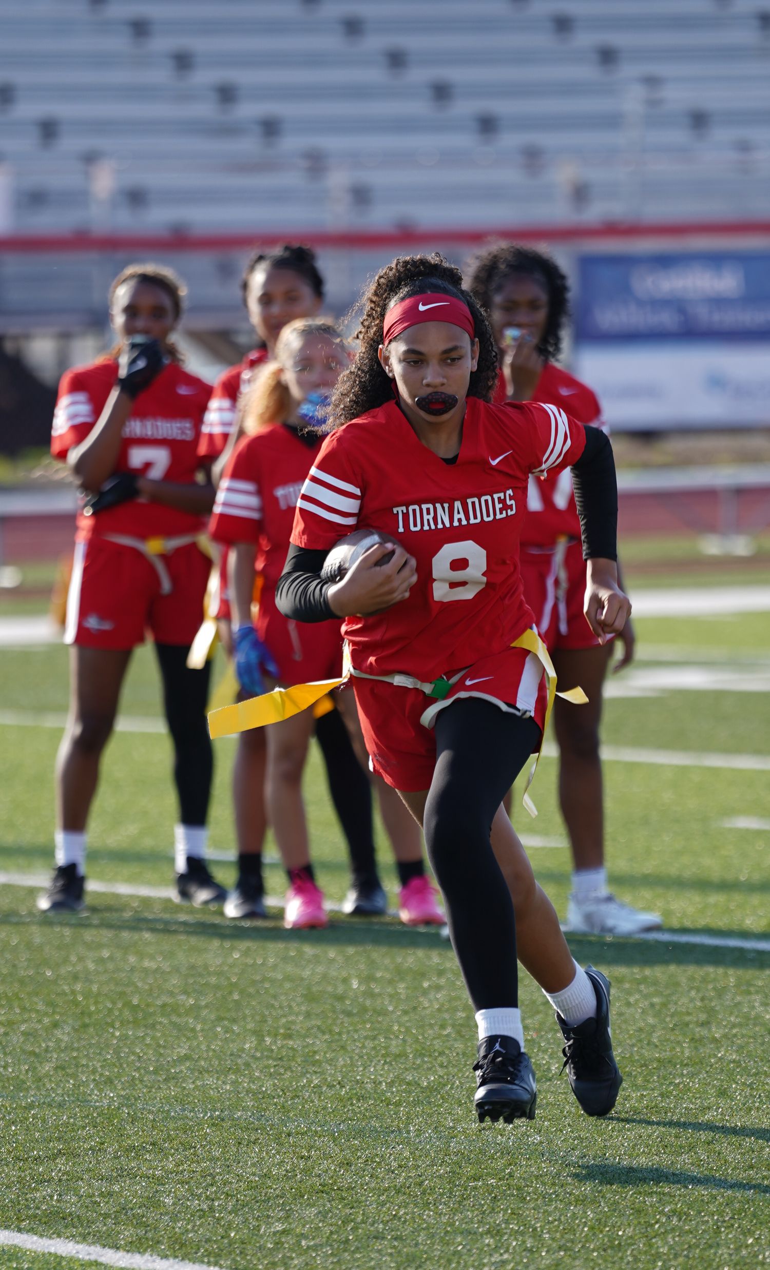 Female flag football player in red Tornadoes jersey #8 runs with ball during game on turf field.
