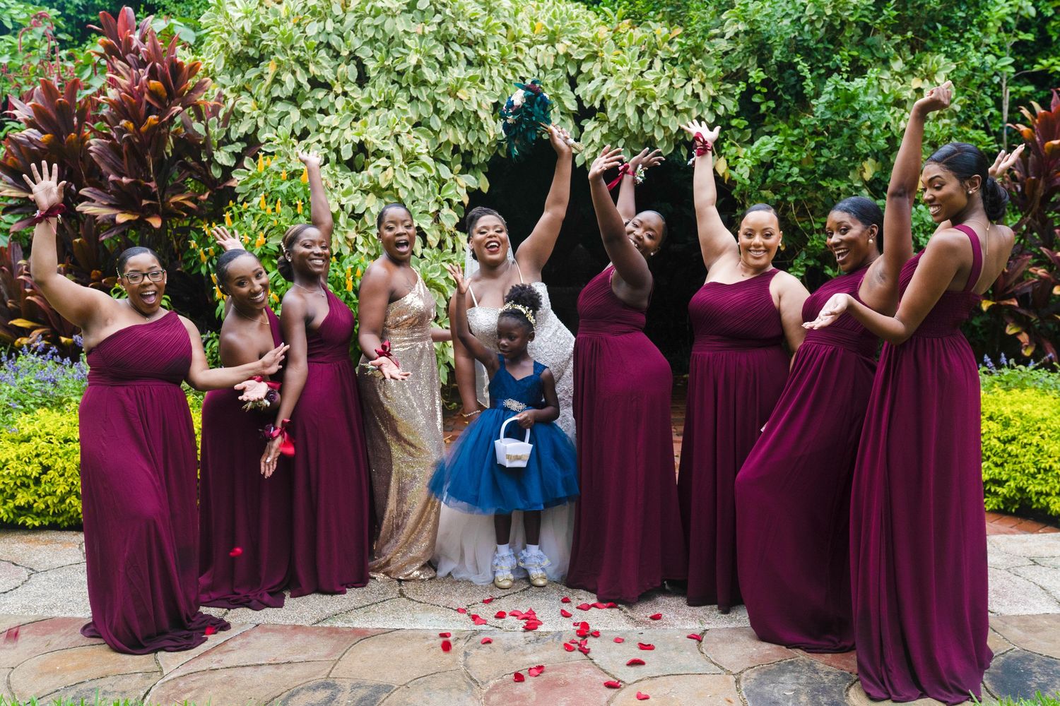 Bride with bridesmaids in burgundy gowns and flower girl in blue dress celebrating outdoors in a lush garden.