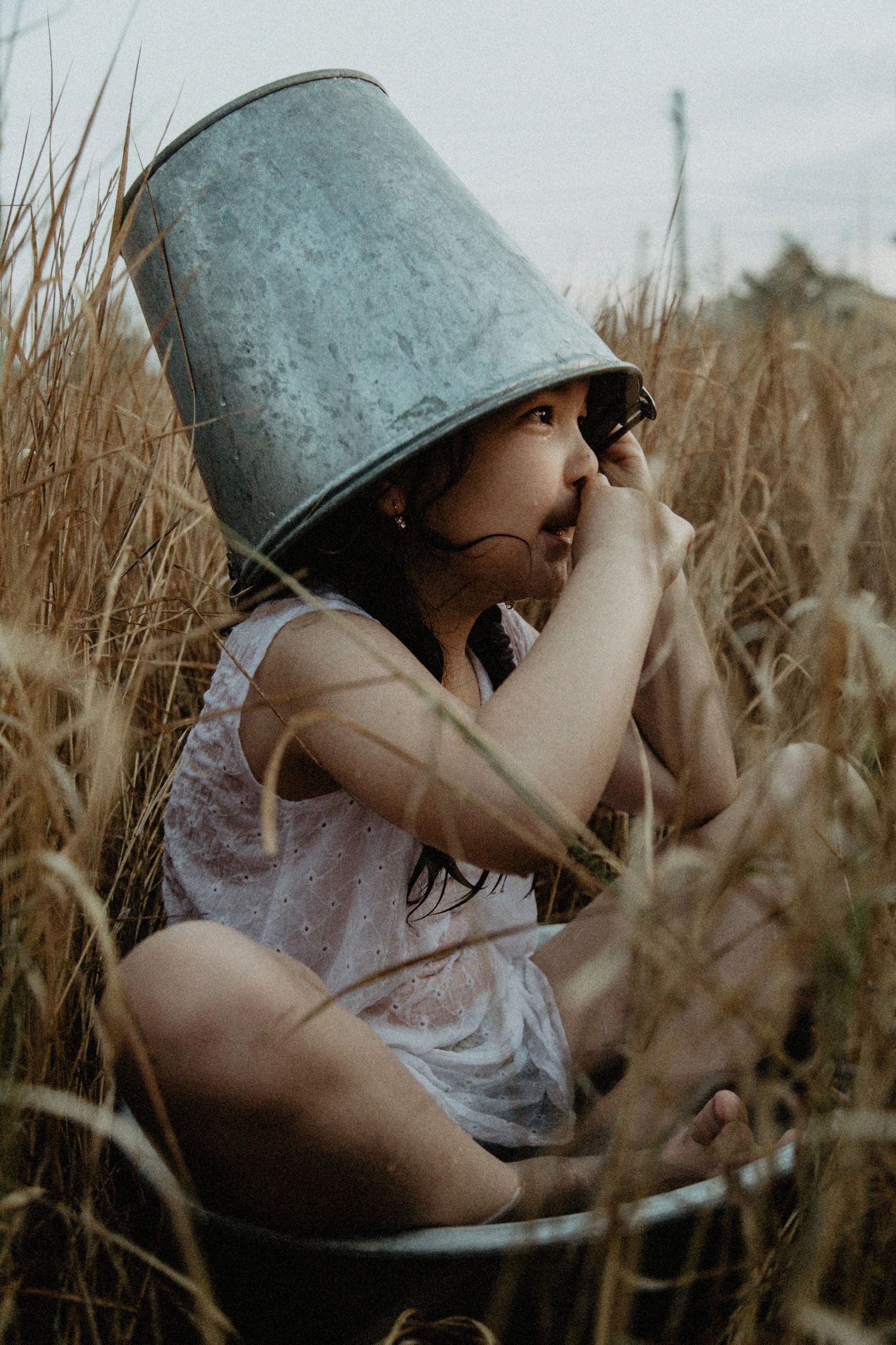A person wearing a metal bucket on their head sits in a wheat field at sunset.