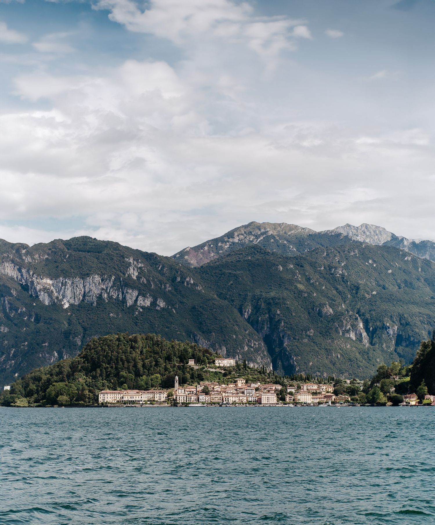 Scenic view of an Italian lakeside village on Lake Como with lush mountains and cloudy sky in the background.
