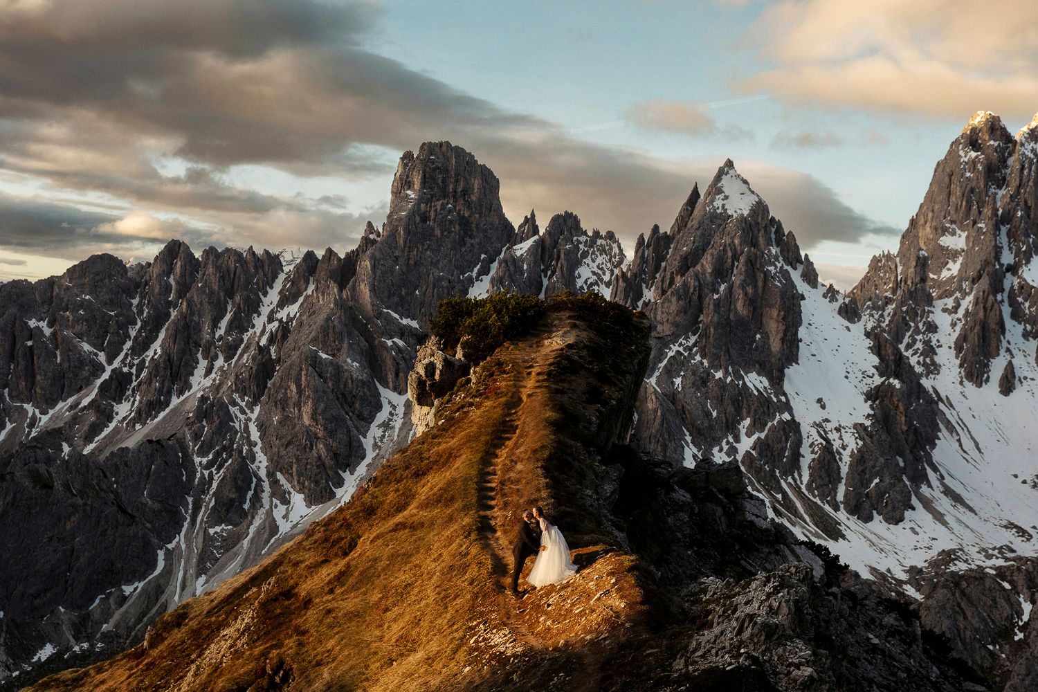 Dramatic mountain landscape at sunset with jagged snow-capped peaks and golden-lit ridge in the foreground.