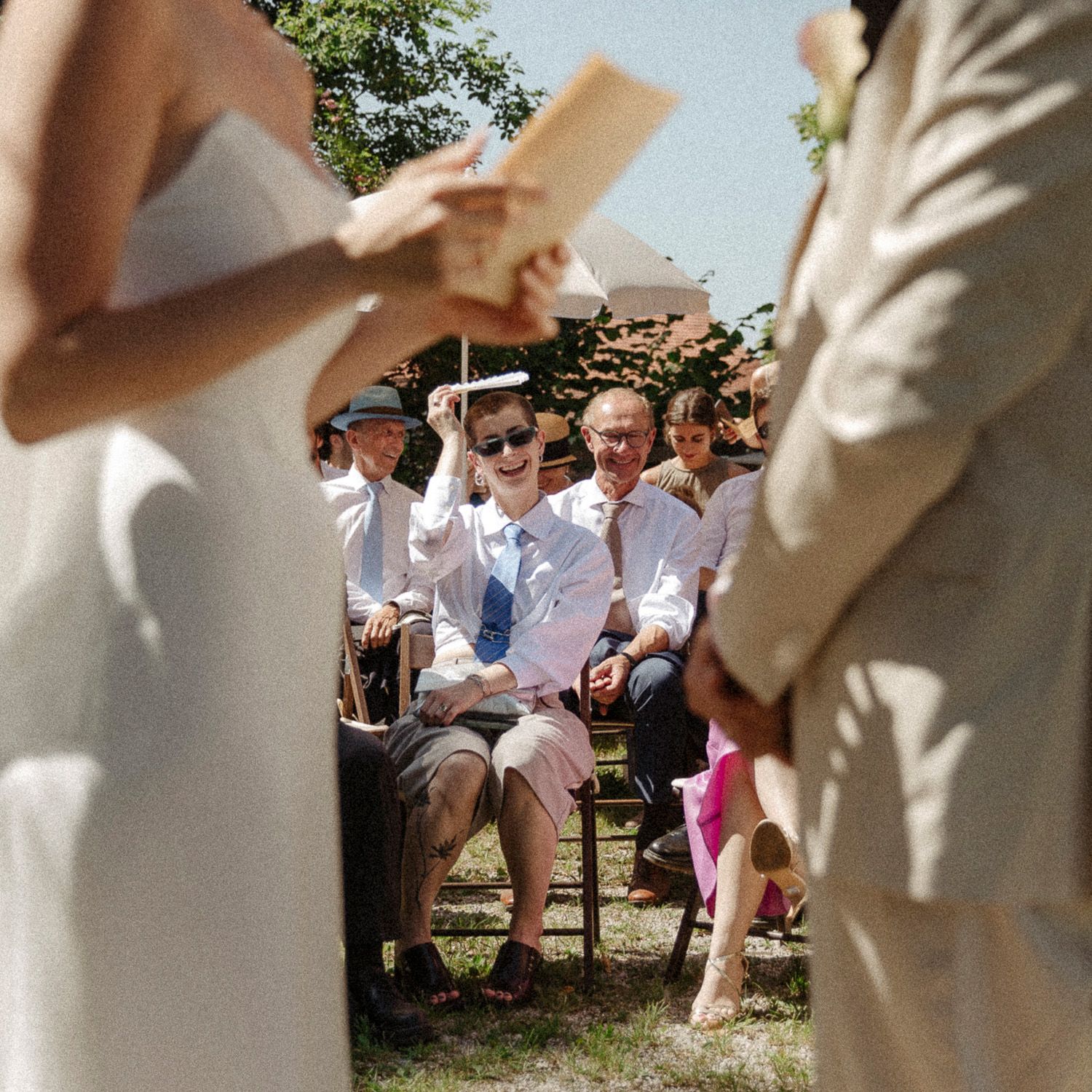 Wedding guests sitting on wooden benches outdoors during a sunny summer ceremony.