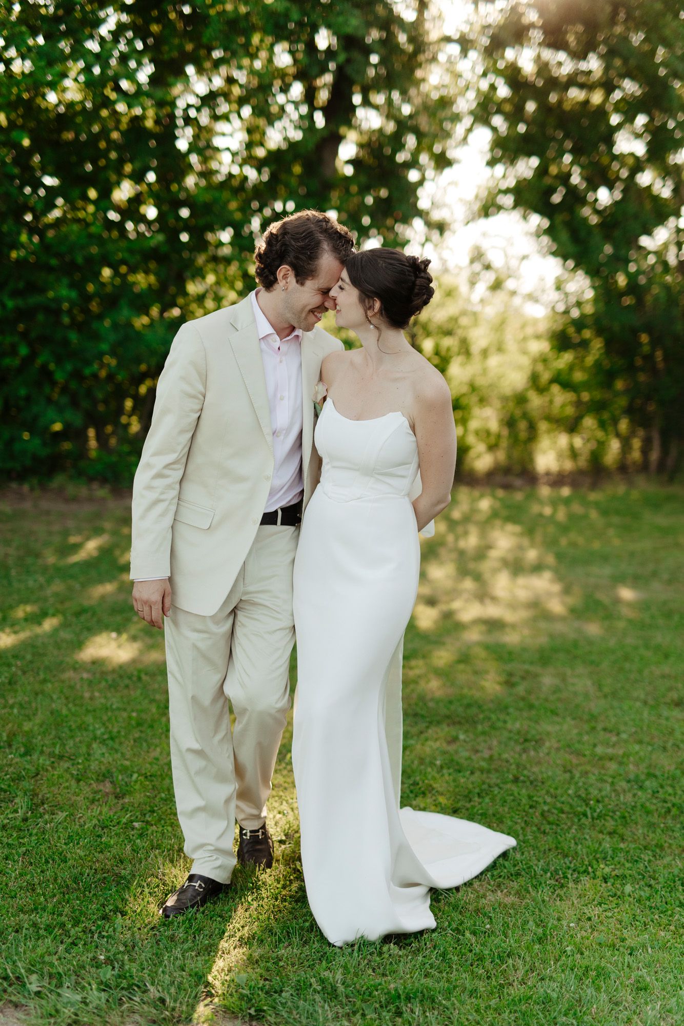 A couple in white wedding attire share a tender moment on a sunlit grassy lawn.