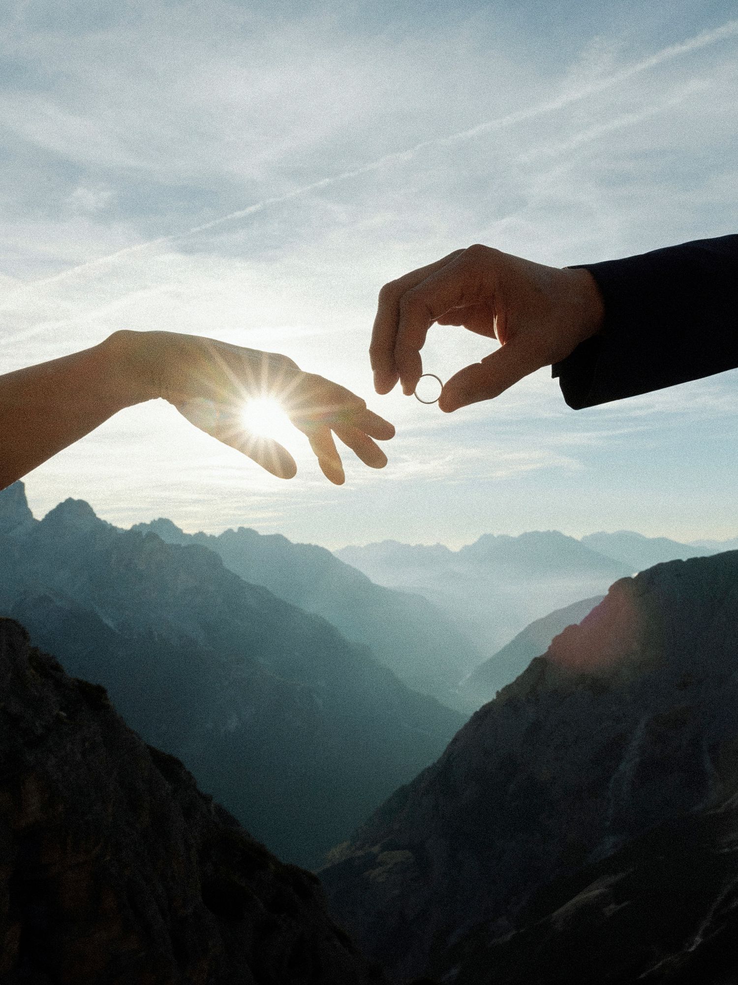 Two hands reaching toward each other over mountain peaks at sunset, with a wedding ring being passed between them.