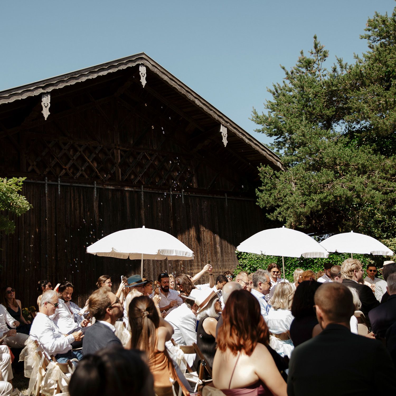Guests gather under white umbrellas outside a rustic wooden barn during an outdoor event.