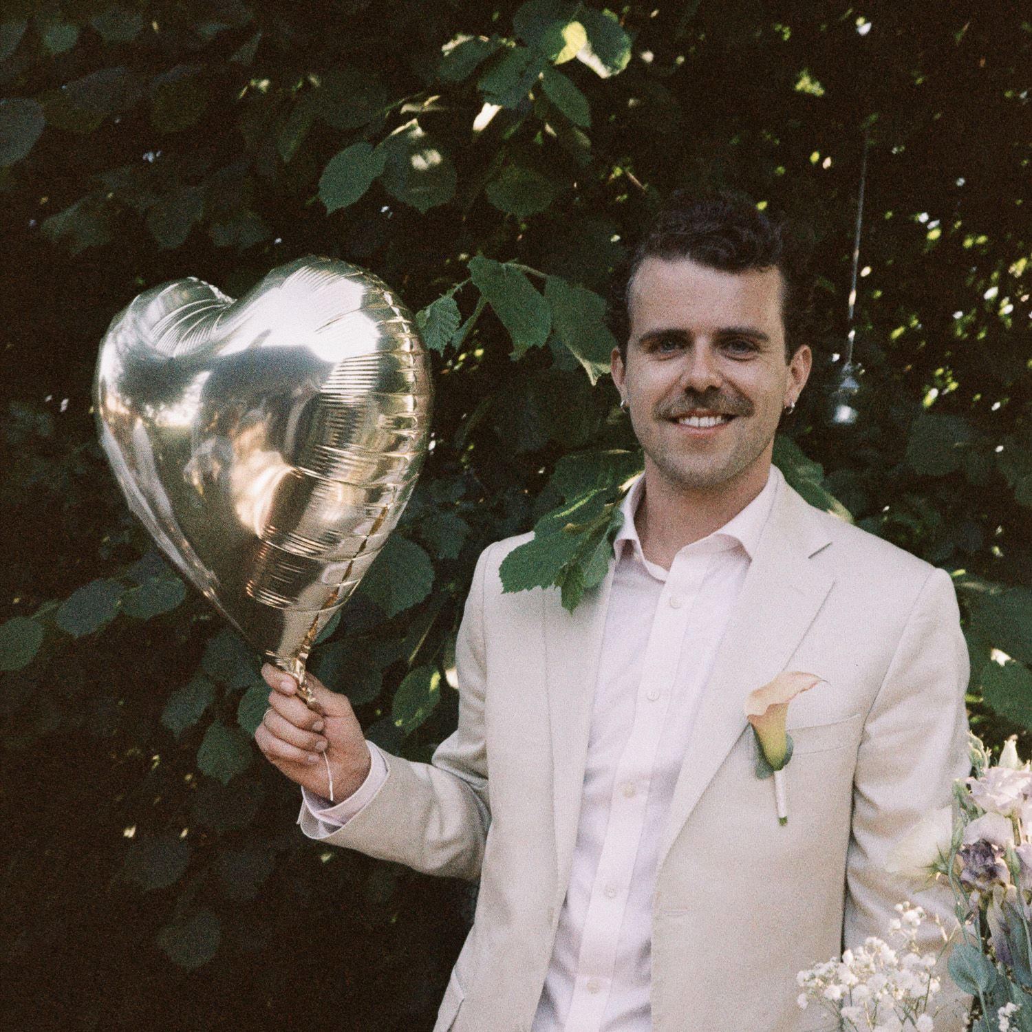 Person in white suit and mustache holds up champagne glasses in garden setting.