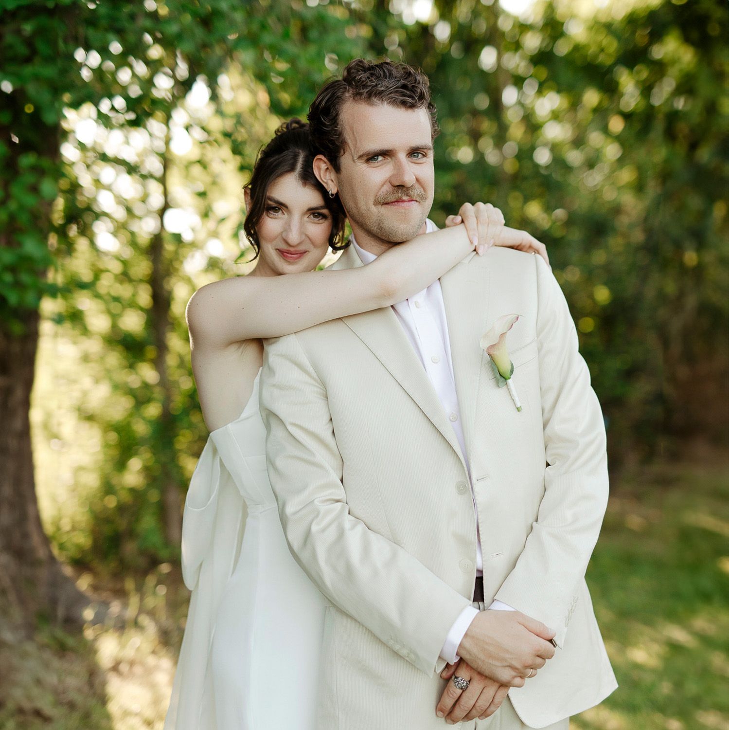 Romantic wedding couple embraces outdoors in natural sunlit garden setting wearing elegant cream colored attire.