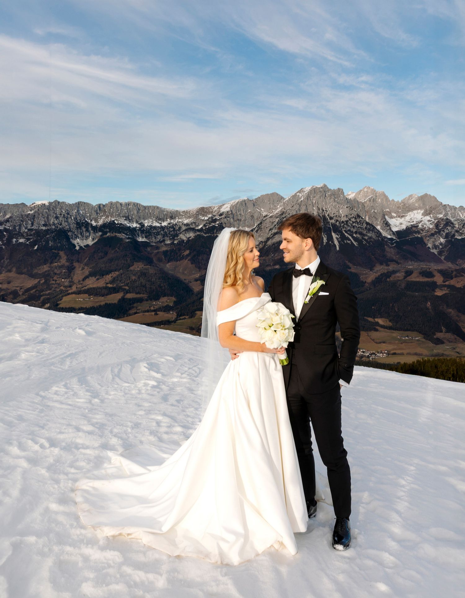 A wedding couple poses for romantic photos on a snowy mountain ridge with dramatic alpine peaks in the background.