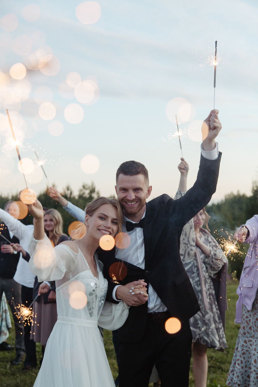 Newlyweds celebrate with sparklers surrounded by guests in an outdoor evening celebration.