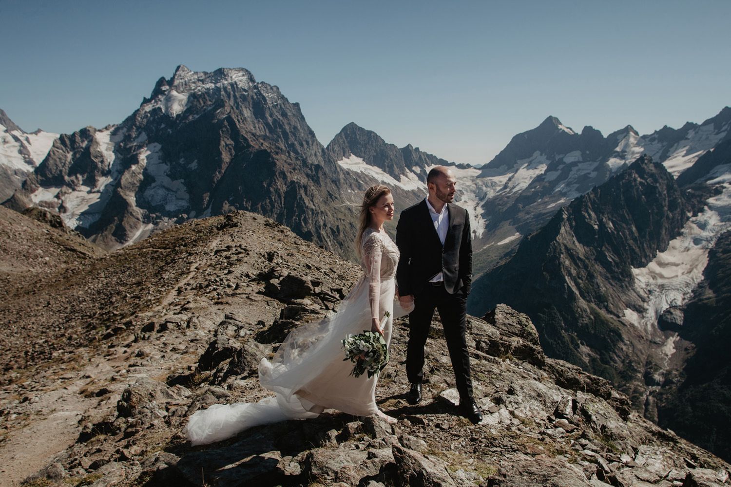 A couple in wedding attire stands atop a mountain peak with snow-capped mountains in the background.