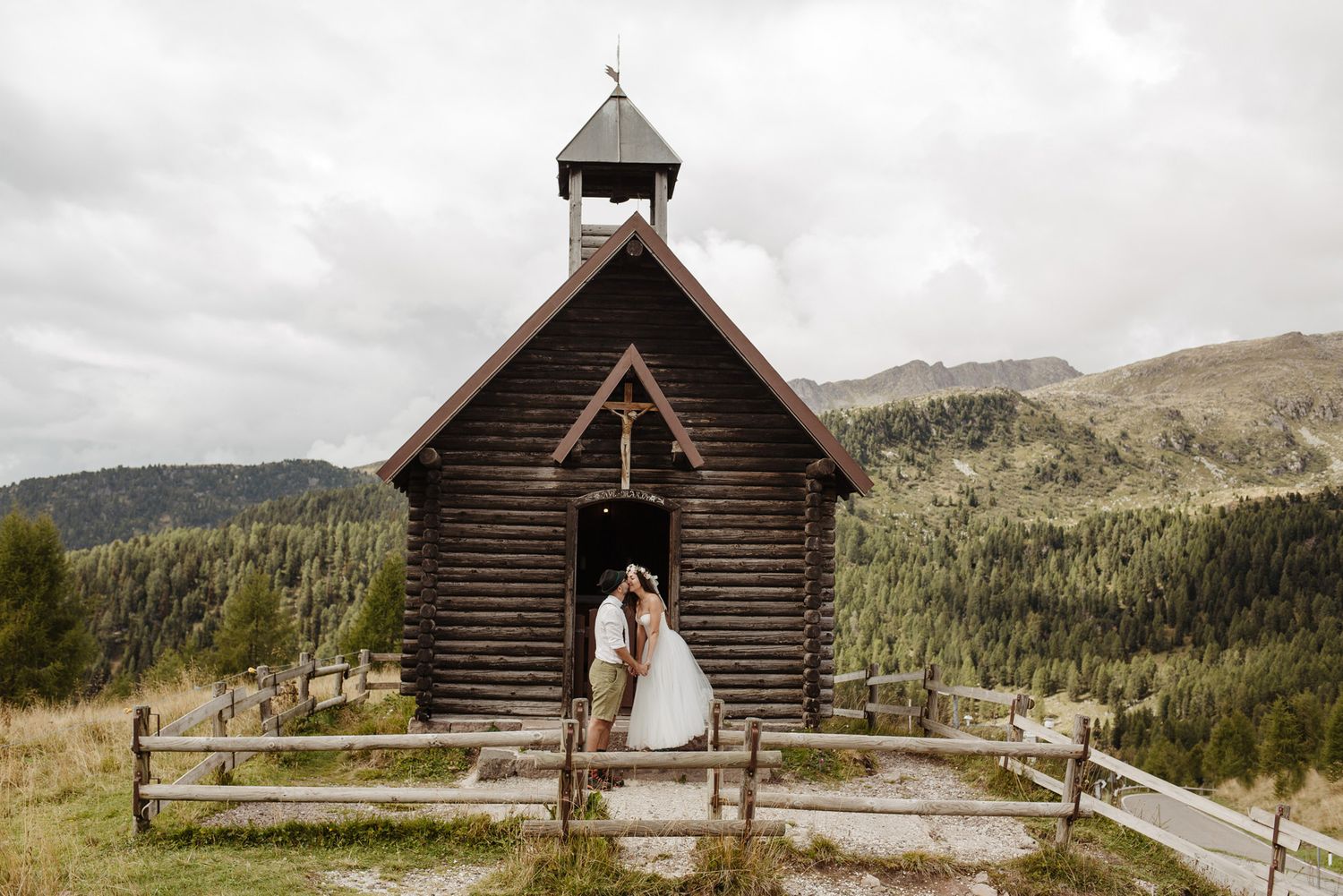 A rustic wooden chapel sits atop a mountain ridge with dramatic mountain peaks and evergreen forests in the background.