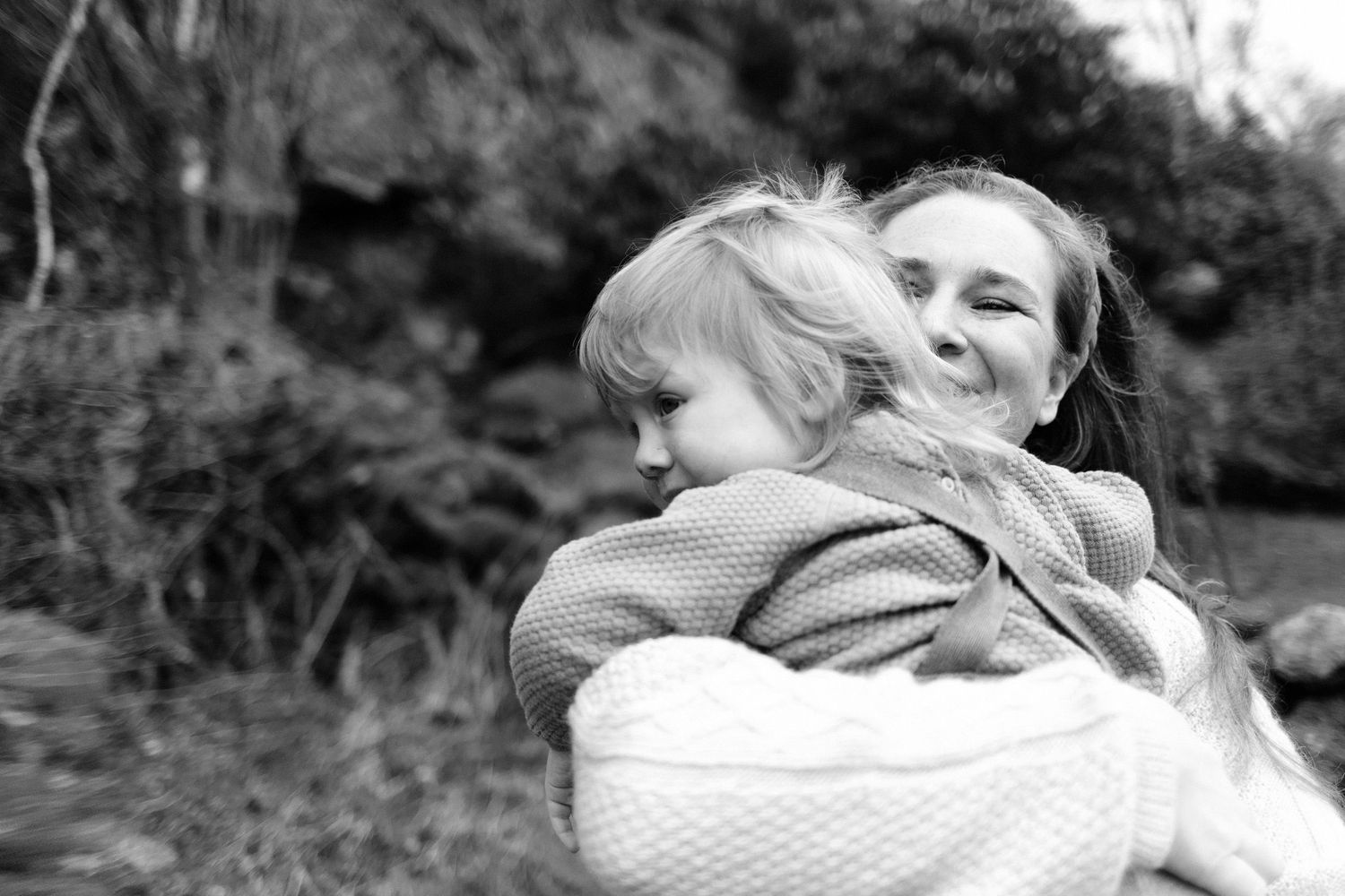 Black and white portrait of a tender embrace between parent and child wrapped in a knit blanket.