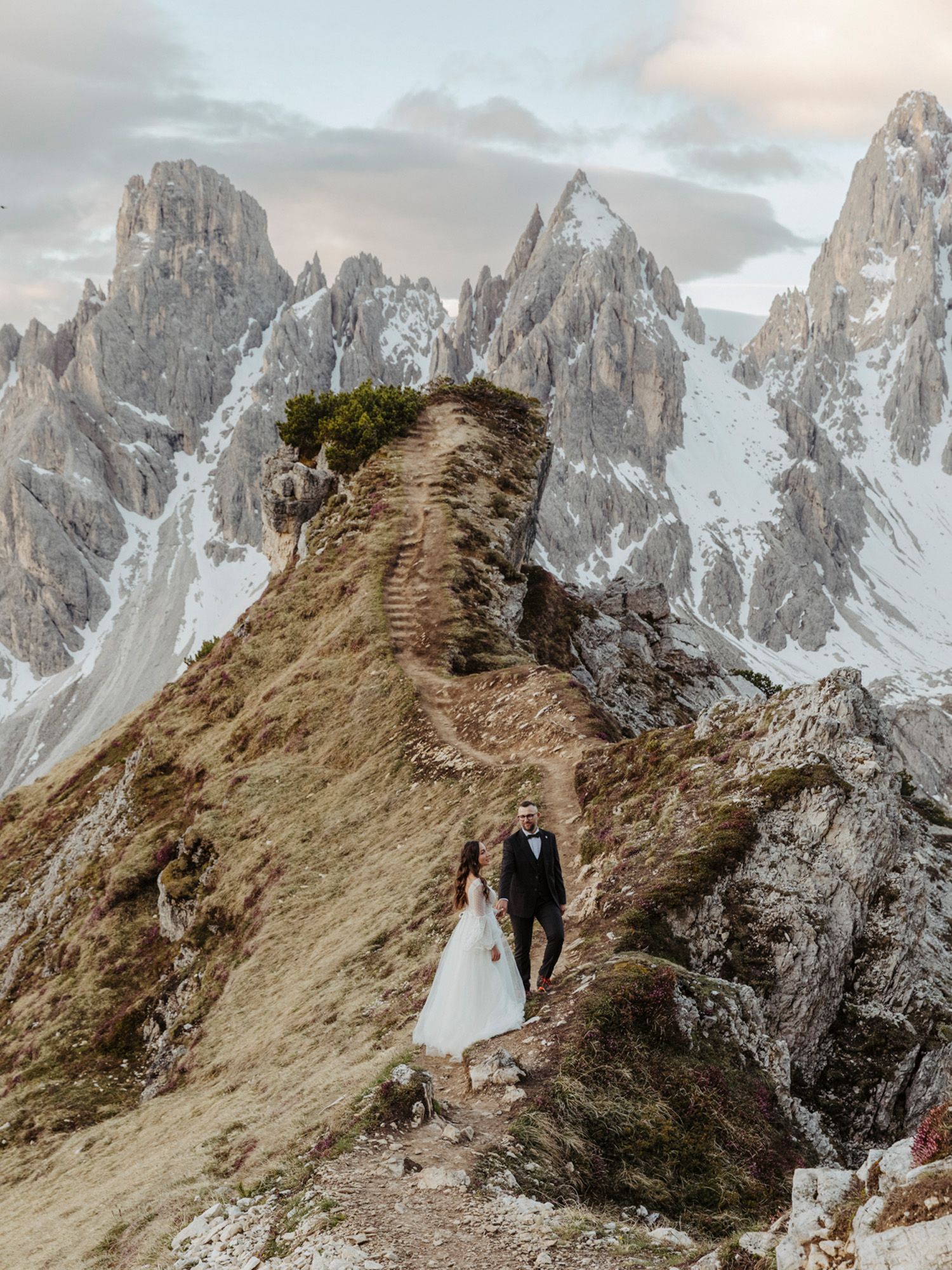 Couple stands on dramatic mountain ridge with jagged Dolomite peaks towering in background during alpine photo session.