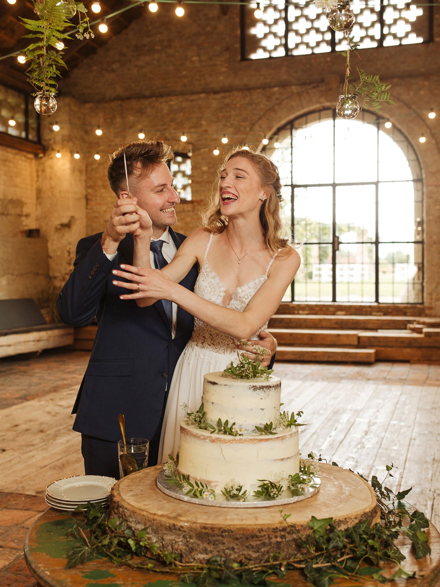Newlyweds share a playful moment while cutting their elegant white wedding cake decorated with greenery in a rustic venue.